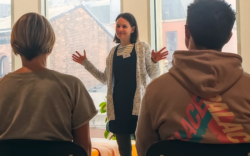 Woman smiling and gesturing confidently while speaking in front of a small audience during a public speaking workshop